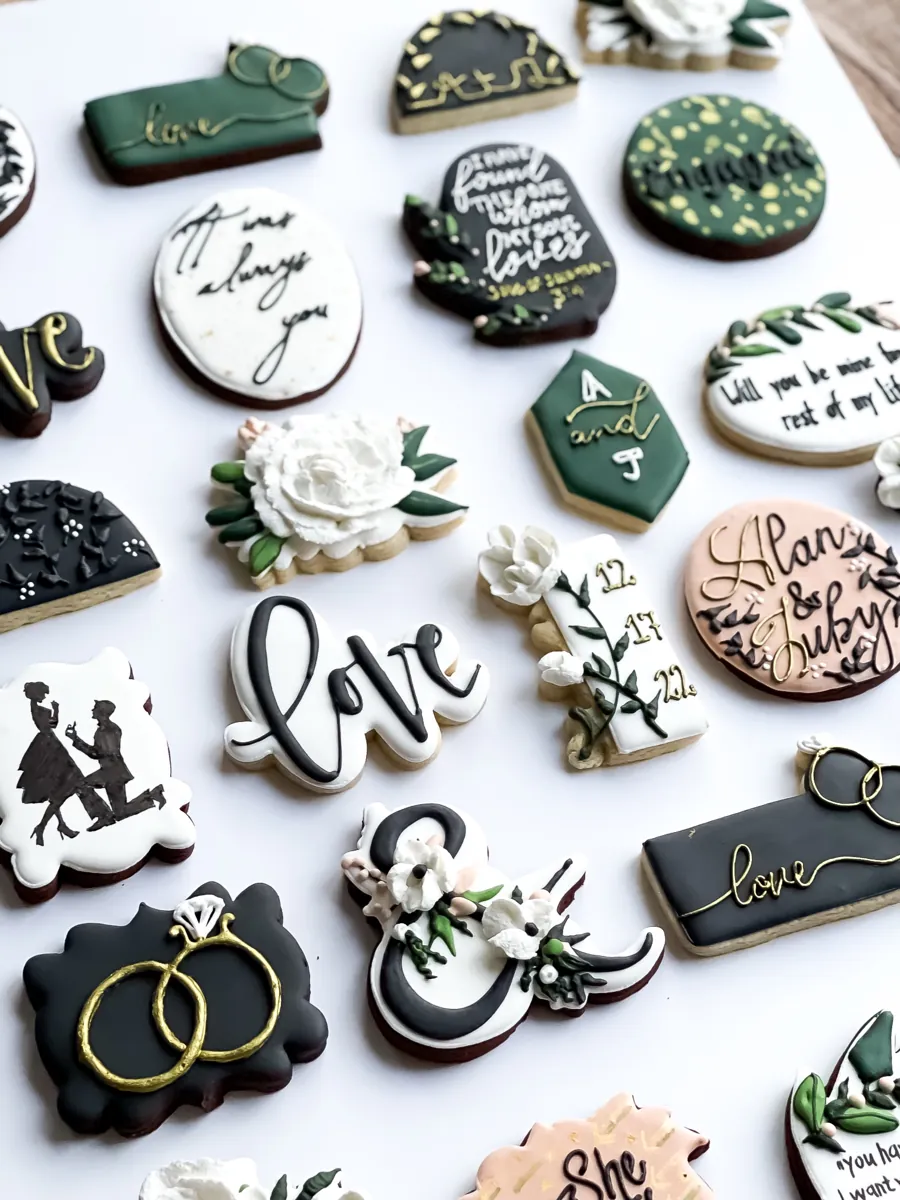 Green, black and white engagement announcement cookies with gold lettering, white flowers and rings
