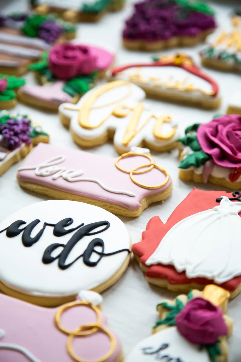 Pink, white and gold bridal cookies with I do lettering, love and wedding rings, wedding dress on a hanger and burgundy buttercream roses