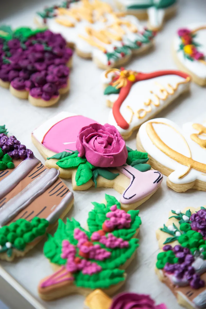 Wine themed bridal shower cookies with buttercream grapes, a pink rose on a wine glass, wine barrel and gold bride lettering on a hanger